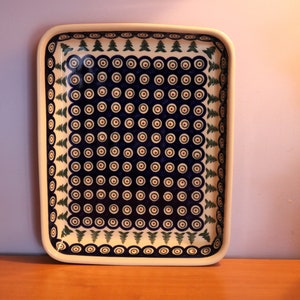 May include: A rectangular ceramic baking dish with a white background and a blue and green pattern. The pattern features a repeating design of circles with a red center and green pine trees.