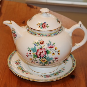 May include: A white teapot with a floral design and gold trim. The teapot has a lid and a handle. It is sitting on a matching saucer.