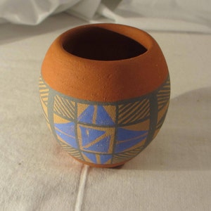 May include: A small, terracotta pot with a geometric design in shades of blue, brown, and tan. The pot is decorated with a central star-shaped pattern.