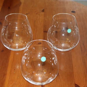 May include: Three clear glass wine glasses on a wooden table. The glasses are round and have a wide base.