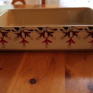 May include: Rectangular beige ceramic baking dish with a floral design. The dish has a cream-colored base with a repeating pattern of red and purple flowers with green leaves along the rim. The dish is sitting on a wooden table.