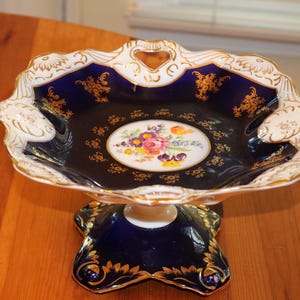 May include: A cobalt blue and gold porcelain serving bowl with a floral design. The bowl has a scalloped edge and a pedestal base.