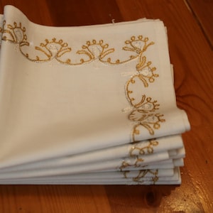 May include: A stack of white linen napkins with gold embroidered floral designs along the edges. The napkins are neatly folded and stacked on a wooden surface, showcasing the decorative trim.