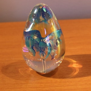 May include: A clear glass paperweight with a blue and green swirl design inside. The paperweight is shaped like an egg and has a smooth, rounded surface.