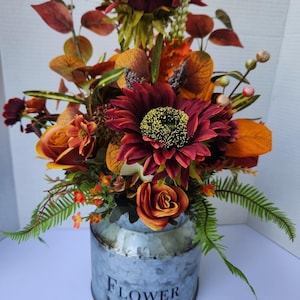 May include: A floral arrangement featuring burgundy sunflowers, orange roses, and various greenery. The flowers are arranged in a galvanized metal container with the text "FLOWER MARKET" in black. The arrangement has a rustic, autumnal theme.