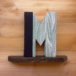 Puede incluir: Una estantería de madera con una letra M en forma de libro hecha de un libro con una cubierta de mármol verde y blanco y una columna vertebral negra. La estantería es de madera oscura.