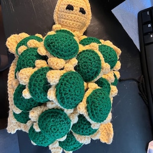 Può includere: Un giocattolo di tartaruga all'uncinetto fatto di filato verde e beige. La tartaruga ha una testa grande e molte tartarughe più piccole impilate sopra.