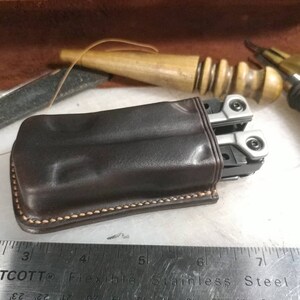 May include: A brown leather case with a stitched edge, designed to hold a multi-tool. The multi-tool is partially visible, with silver metal parts and a black handle.