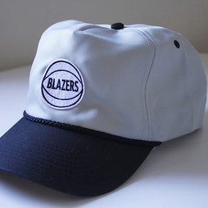 May include: A white and black baseball cap with a navy blue embroidered patch that says "BLAZERS" with a basketball graphic.