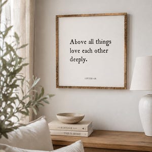 May include: A square wall sign with a natural wood frame and a white background featuring black text that reads: Above all things love each other deeply. 1 PETER 4:8. The sign is mounted on a plain wall above a wooden surface with books and a bowl.