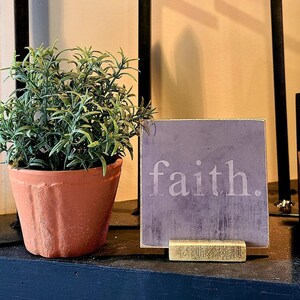May include: A terracotta-colored potted plant with green foliage sits next to a small, square sign that reads "faith." in white lettering. The sign is propped up by a small, gold-colored stand. Black metal crosses are in the background.