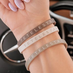 May include: Three silicone wristbands in varying shades of beige and brown, each featuring the word "TETELESTAI" embossed in white capital letters. The bracelets are worn stacked together on a wrist, showcasing a smooth, matte finish.