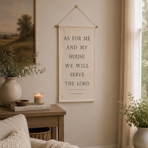 May include: A rectangular off-white fabric wall hanging suspended from a wooden dowel with a thin cord. The text printed in black reads: AS FOR ME AND MY HOUSE WE WILL SERVE THE LORD, followed by a horizontal line and the citation JOSHUA 24:15.