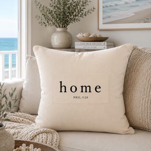 May include: A square off-white throw pillow featuring a rectangular patch in the center. The patch displays the word "home" in a black serif font with the text "PHIL.3:20" printed in a smaller black font directly underneath the word.