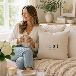 May include: A square beige throw pillow featuring a rectangular fabric patch centered on the front. The patch displays the word rest in a black serif font, with the text HEBREWS 4:1 printed in a smaller black font directly underneath it.