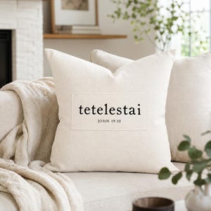 May include: A square off-white throw pillow featuring a rectangular fabric patch centered on the front. The patch displays the black printed text "tetelestai" in a serif font, with the smaller text "JOHN 19:30" positioned directly underneath.