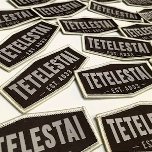 May include: Multiple black and white fabric patches with the text "TETELESTAI EST. AD33" in a white font. The patches are rectangular with rounded corners and have a white border.