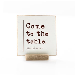 May include: White wooden sign with the text "Come to the table." in brown lettering. The sign also includes the text "REVELATION 19:9" in smaller lettering. The sign is standing on a small wooden stand.