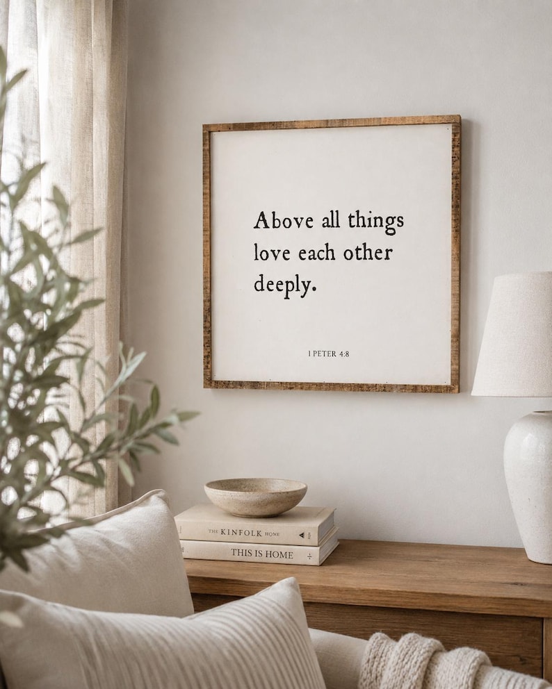 May include: A square wall sign with a natural wood frame and a white background featuring black text that reads: Above all things love each other deeply. 1 PETER 4:8. The sign is mounted on a plain wall above a wooden surface with books and a bowl.