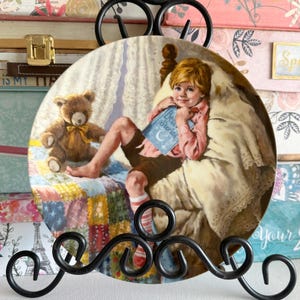May include: Decorative plate featuring a child reading "Dear Stories" next to a teddy bear. The plate has a detailed illustration with a warm color palette, set against a background of a bed with a patchwork quilt. The plate is held by a black metal stand.