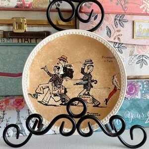 May include: A decorative plate with a cream-colored rim and a tan center featuring a vintage illustration of people walking. The plate has the text "Promenade à Paris" and "Vote for Al". The plate is displayed on a black metal stand.