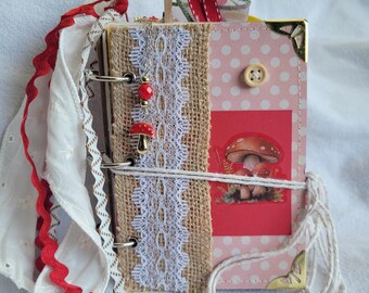 Könnte beinhalten: Ein rosa und rotes Journal mit einem Pilz-Design, verziert mit Sackleinen, Spitze und rot-weißem Band. Das Journal hat eine goldene Ecke und einen Knopf.