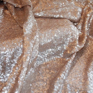 May include: A close-up of a fabric with a shimmering, iridescent finish. The fabric is made of sequins in shades of brown and silver, creating a luxurious and glamorous look.