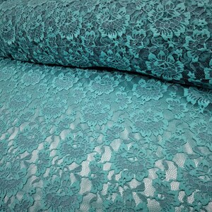 Pode incluir: Um rolo de tecido de renda na cor verde-azulado com um padr&atilde;o floral detalhado. O design intrincado consiste em flores e folhas interconectadas, criando uma textura delicada e vazada por todo o material. O tecido est&aacute; dobrado sobre si mesmo.