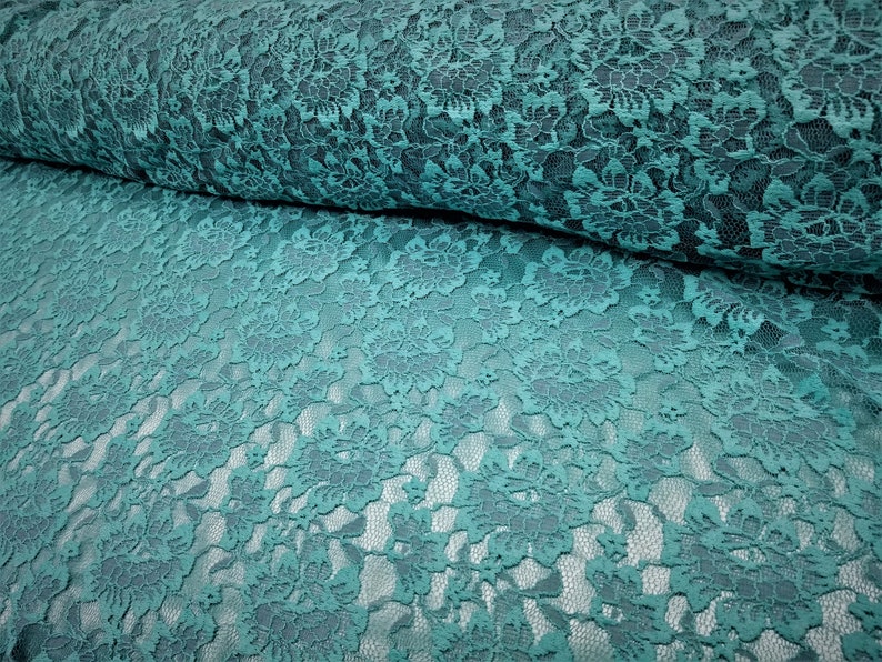 Pode incluir: Um rolo de tecido de renda na cor verde-azulado com um padr&atilde;o floral detalhado. O design intrincado consiste em flores e folhas interconectadas, criando uma textura delicada e vazada por todo o material. O tecido est&aacute; dobrado sobre si mesmo.