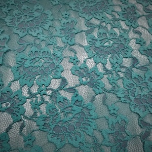 Pode incluir: Um tecido de renda verde-azulado com um padr&atilde;o floral denso e intrincado, bordas recortadas e um fundo de malha fina. O material apresenta um design bot&acirc;nico consistente e repetitivo, caracterizado por p&eacute;talas e folhas interligadas.