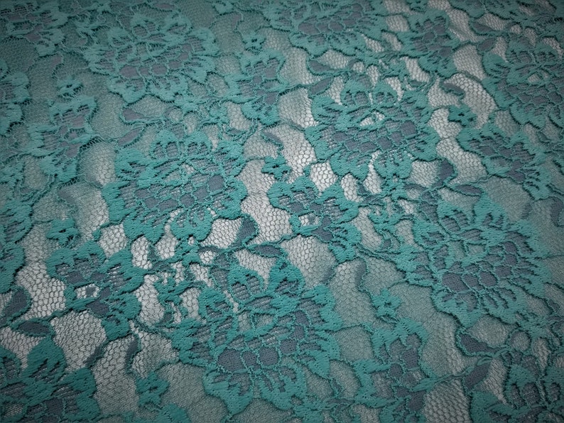 Pode incluir: Um tecido de renda verde-azulado com um padr&atilde;o floral denso e intrincado, bordas recortadas e um fundo de malha fina. O material apresenta um design bot&acirc;nico consistente e repetitivo, caracterizado por p&eacute;talas e folhas interligadas.