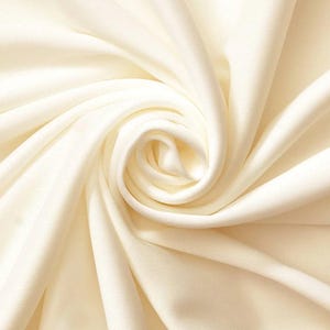 May include: Close-up of a smooth, ivory-colored fabric with a soft, flowing texture. The fabric is draped and folded, creating a swirling pattern.