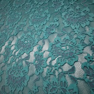 Pode incluir: Um tecido de renda na cor verde-azulado com um padr&atilde;o floral denso e complexo de flores e folhas interligadas. O material tem um fundo de malha semitransparente, criando um efeito visual em camadas com o bordado opaco das flores.