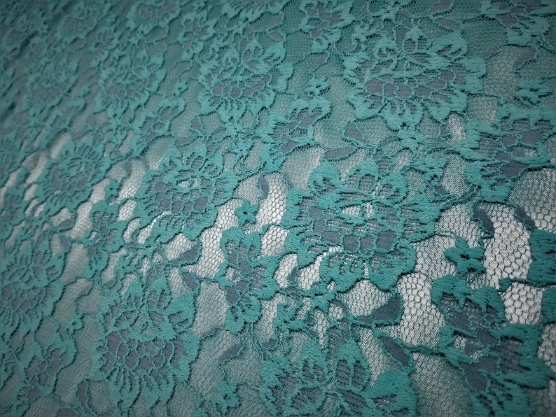 Pode incluir: Um tecido de renda na cor verde-azulado com um padr&atilde;o floral denso e complexo de flores e folhas interligadas. O material tem um fundo de malha semitransparente, criando um efeito visual em camadas com o bordado opaco das flores.