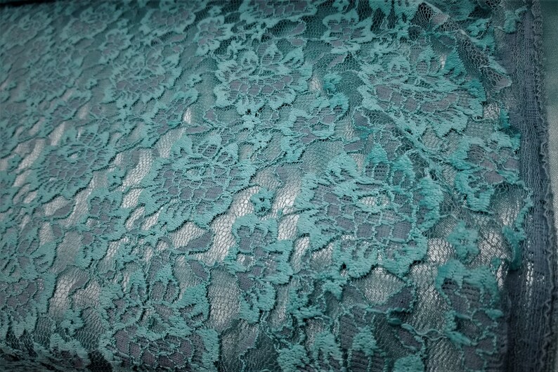 Pode incluir: Um tecido de renda na cor verde-azulado com um padr&atilde;o floral intrincado e bordas festonadas. O tecido delicado exibe um design bot&acirc;nico repetitivo com detalhes vazados, apresentando uma textura consistente e um tom azul-esverdeado vibrante.