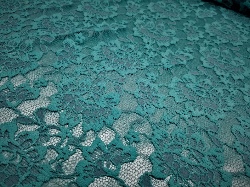 Pode incluir: Um tecido de renda verde-azulado com um padr&atilde;o floral repetitivo, bordas recortadas intrincadas e um fundo de malha fina. O material exibe um tom azul-esverdeado profundo e consistente, destacando o bordado detalhado das flores.
