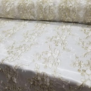 Pode incluir: Rolo de tecido de renda marfim com um padrão floral. A renda apresenta bordados florais em relevo na cor dourada. O tecido é drapeado sobre uma superfície, mostrando sua textura e design.