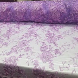 May include: A roll of lavender floral lace fabric. The fabric features an intricate embroidered design of flowers and leaves. The lace is a soft, sheer material, perfect for dresses, veils, or other craft projects.