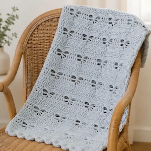 May include: A light blue crochet blanket featuring a repeating pattern of small, stylised dragonfly motifs arranged in horizontal rows. The blanket has a scalloped edge border and is crafted with a textured, openwork stitch design throughout.