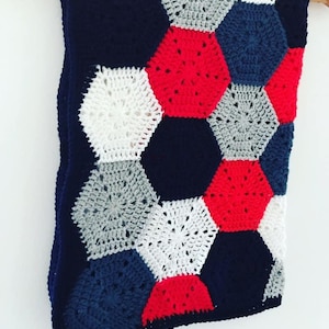 May include: A crocheted blanket with a geometric pattern in shades of navy blue, red, white, and gray. The blanket is made of hexagonal shapes and is hanging on a hanger.