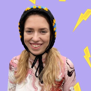 May include: A woman wearing a black crocheted hat with yellow and blue accents. The hat has a long black crocheted tie. The woman is wearing a pink and brown marbled long-sleeved shirt.