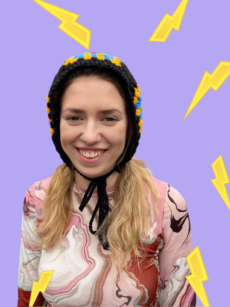 May include: A woman wearing a black crocheted hat with yellow and blue accents. The hat has a long black crocheted tie. The woman is wearing a pink and brown marbled long-sleeved shirt.