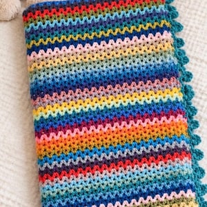 May include: A rectangular crochet blanket featuring a vibrant chevron pattern with horizontal stripes in shades of blue, green, yellow, orange, red, purple, and pink. The blanket is finished with a dark teal scalloped edge border.