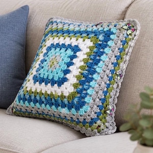Peut inclure: Un coussin carré au crochet présentant un motif de granny square dans des tons de bleu, vert, blanc et gris. Le coussin possède une bordure festonnée grise agrémentée de petites décorations en forme de papillons colorés.