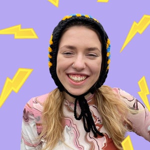 May include: A person wearing a black, yellow, and blue crocheted hat with a black string tie. The hat covers their ears and the person is smiling. The background is a light purple with yellow lightning bolts.