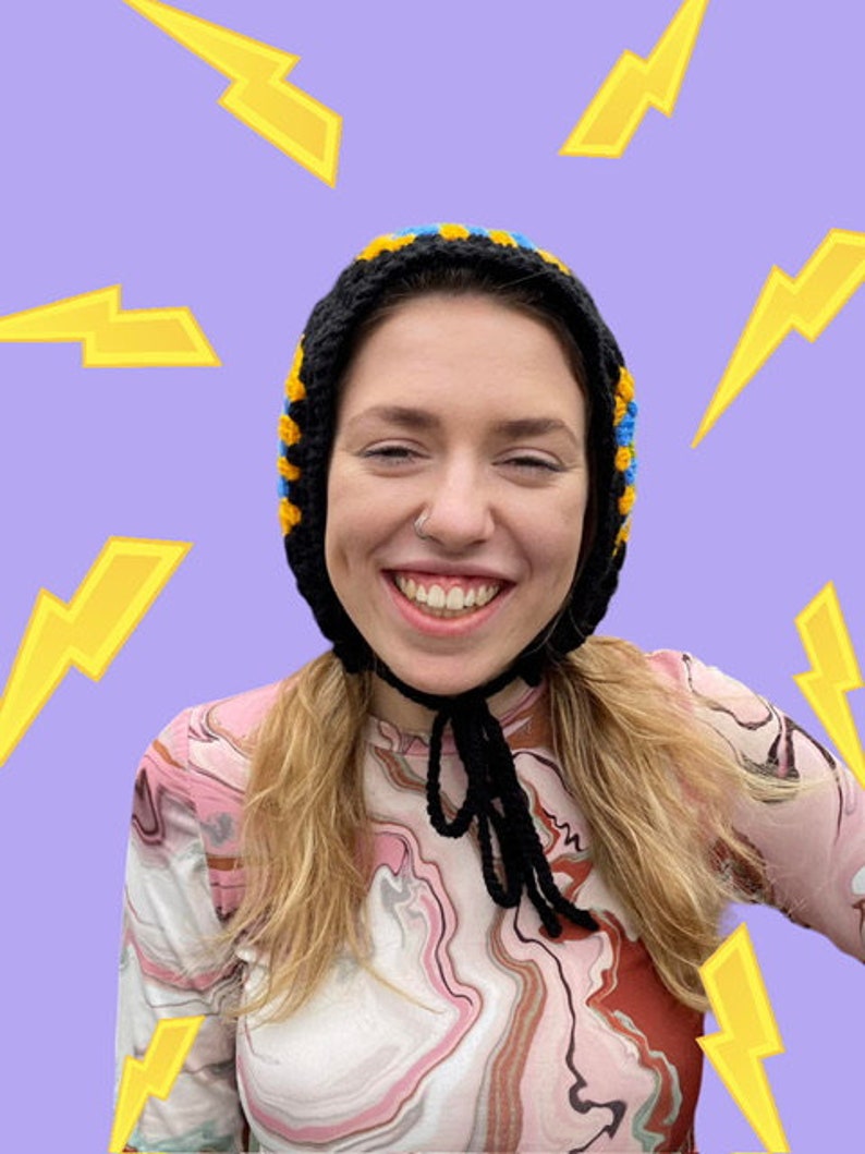 May include: A person wearing a black, yellow, and blue crocheted hat with a black string tie. The hat covers their ears and the person is smiling. The background is a light purple with yellow lightning bolts.