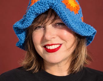 May include: A blue crochet bucket hat featuring a repeating pattern of orange and yellow flower motifs. The hat is constructed with a textured yarn stitch and has a wide, flexible brim that frames the head with a soft, rounded shape.