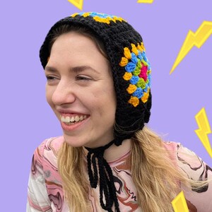 May include: A black crochet hat with a colorful granny square design. The hat has a long tie that can be worn around the neck.