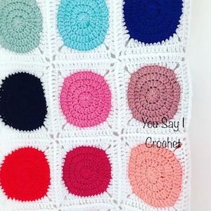 May include: A close-up of a handmade crochet blanket with a repeating pattern of colourful circles on a white background. The circles are in various colours, including blue, pink, and red. The text "You Say I Crochet" is visible.