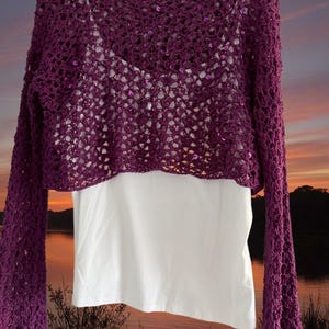 May include: A purple crochet bolero with long sleeves and a delicate open weave. The bolero is layered over a white top. The sleeves are flared at the wrist. Small sequins are scattered across the crochet.